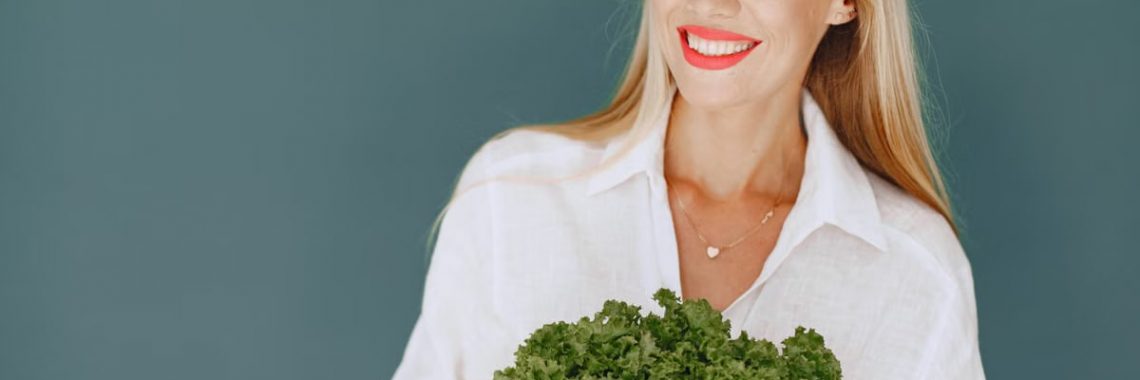 Cheerful woman with blonde hair holding fresh lettuce, promoting healthy living in a studio setting.